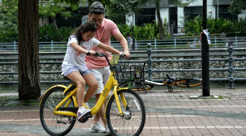 Father teaches daughter to ride a yellow bicycle in a Singapore park. Bonding moment.